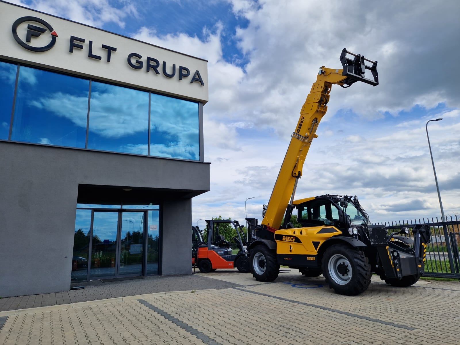 Żółty wózek widłowy Dieci na tle nowoczesnego budynku z logo FLT Grupa i odbijającym się niebem w oknach. W tle widoczny pomarańczowy wózek.