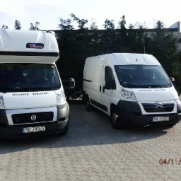 Dwa białe busy, Fiat Ducato z wysoką zabudową i Citroen Jumper, zaparkowane na kostce brukowej przed zielonym żywopłotem. Widoczne tablice rejestracyjne.