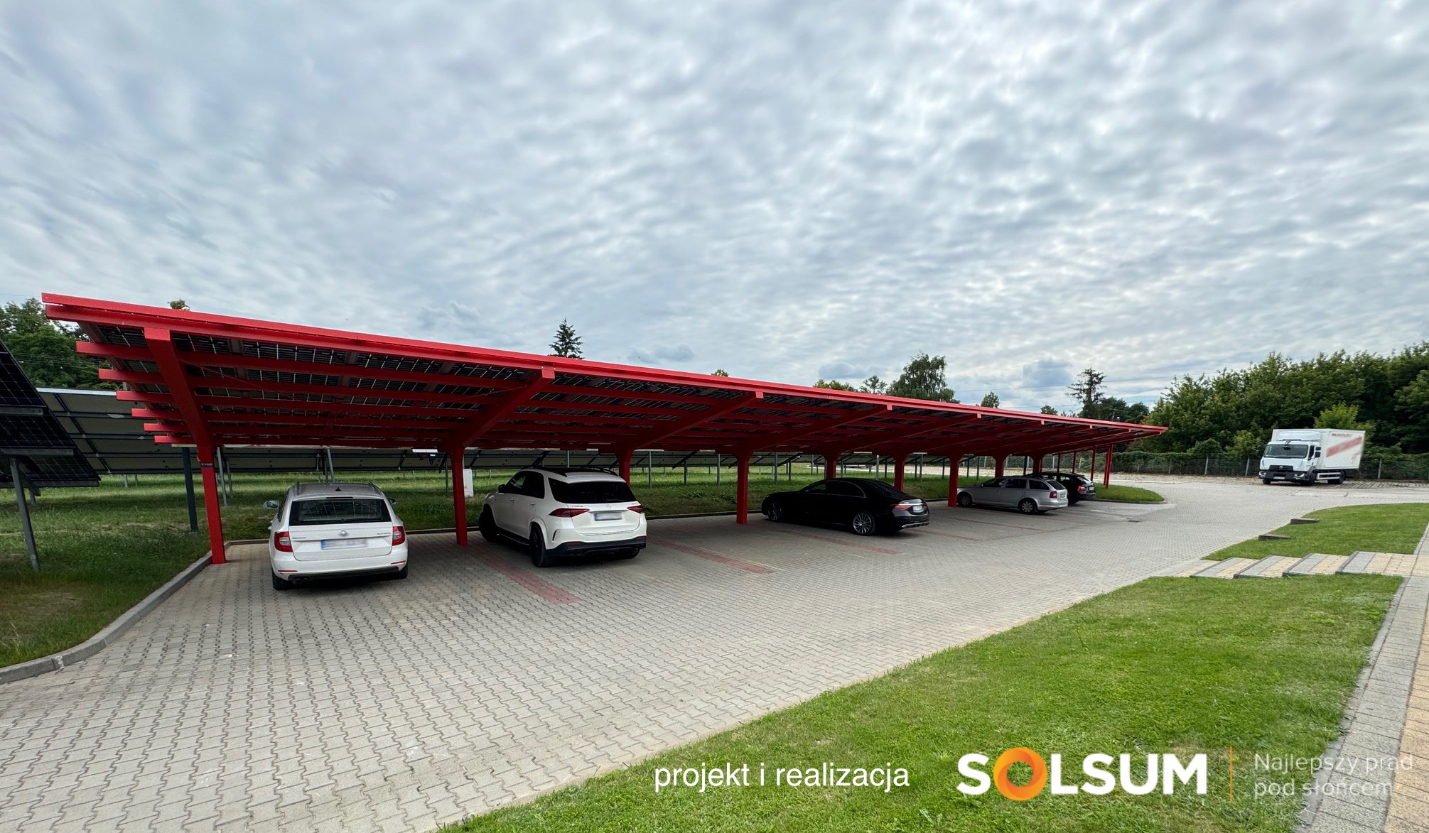 Zadaszony parking z panelami słonecznymi w czerwonej konstrukcji, zaparkowane samochody, widok na zachmurzone niebo i zieleń wokół.