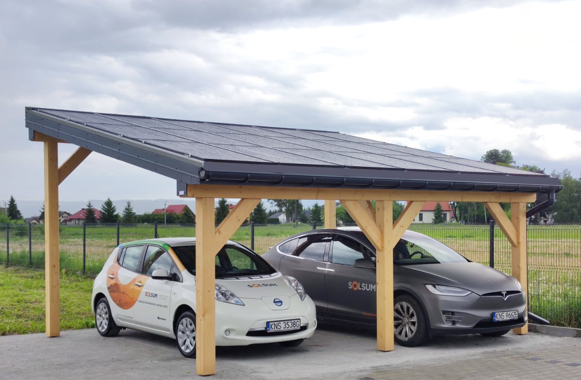 Wiata garażowa z panelami na dachu, pod którą zaparkowane są dwa samochody elektryczne: biały Nissan Leaf z logo firmy i szary Tesla Model X.