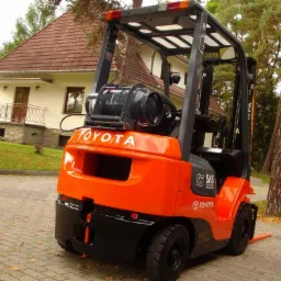 Pomarańczowo-czarny wózek widłowy Toyota 7 FGF 15 z butlą gazową na tle domu z brązowym dachem i zielonym ogrodem.