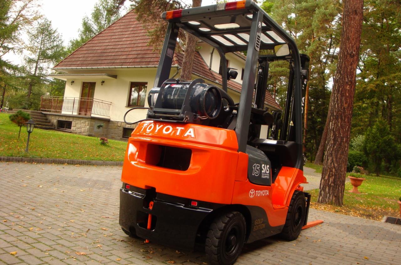 Pomarańczowo-czarny wózek widłowy Toyota 7 FGF 15 z butlą gazową na tle domu z brązowym dachem i zielonym ogrodem.