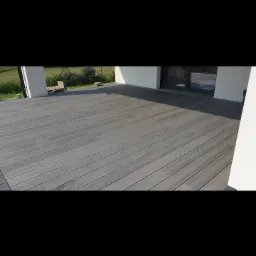 Szara deska tarasowa z kompozytu drewna, widok z góry, częściowo zacieniona, z fragmentem białej ściany budynku w tle.