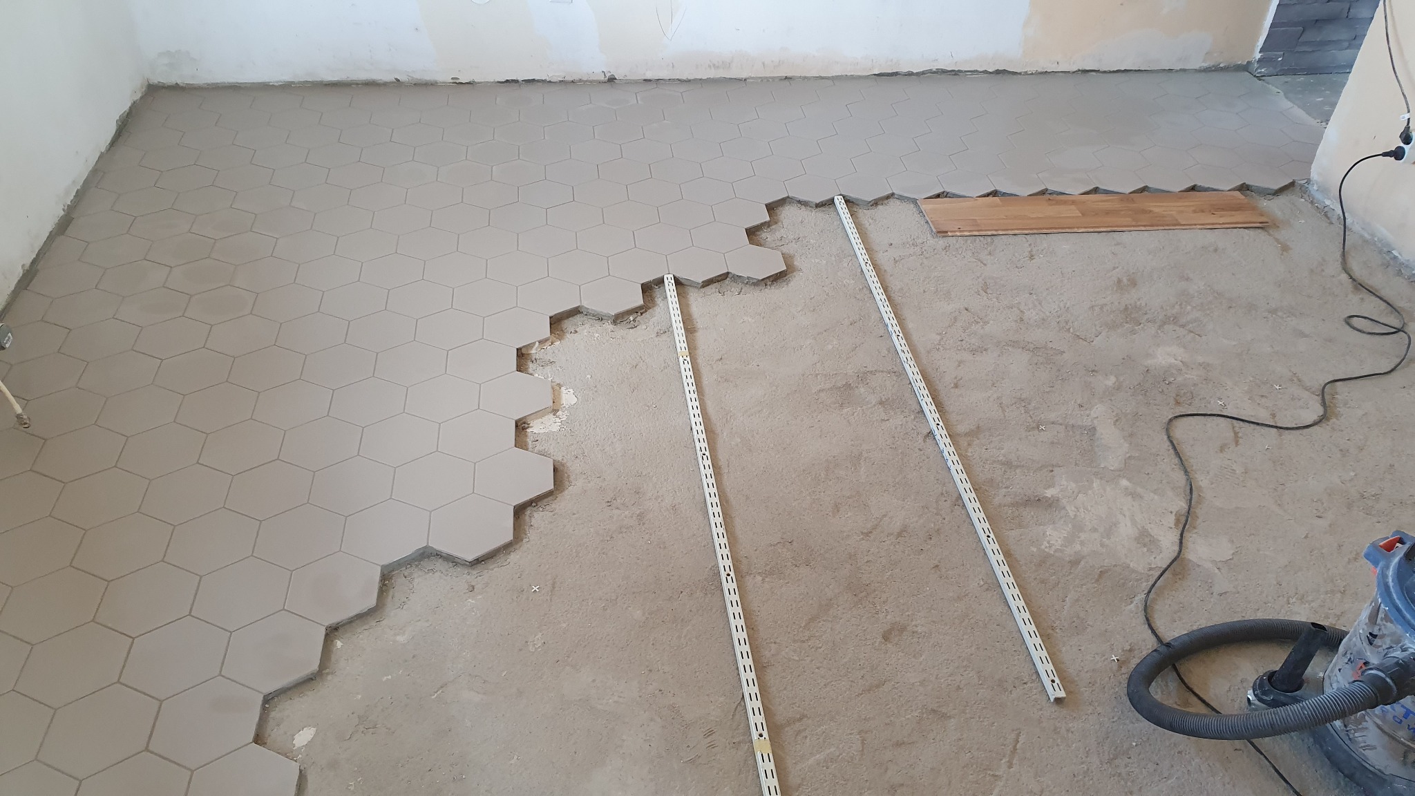 Proces układania heksagonalnych płytek podłogowych w odcieniach szarości, widoczne listwy poziomujące i odkurzacz przemysłowy, fragment surowego podłoża.