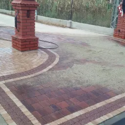 Ułożona kostka brukowa w odcieniach brązu i beżu, z dekoracyjnym wzorem w kształcie łuku, częściowo pokryta piaskiem, otoczona ceglanymi słupkami z czerwonymi daszkami, w tle ogrodzenie z siatki...