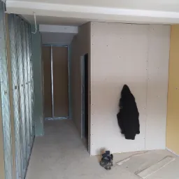 Wnętrze w trakcie montażu ścian działowych z płyt kartonowo-gipsowych, widoczne metalowe profile, płyty KG z widocznymi wkrętami, czarna kurtka wisząca na ścianie, robocze buty i fragmenty płyt...