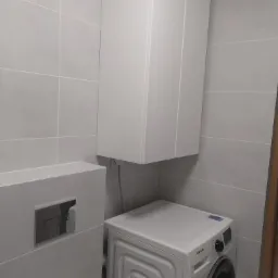 Nowoczesna łazienka z szarymi, kwadratowymi płytkami, zabudowaną toaletą z chromowanym przyciskiem spłukiwania, białą szafką wiszącą i pralką Samsung.