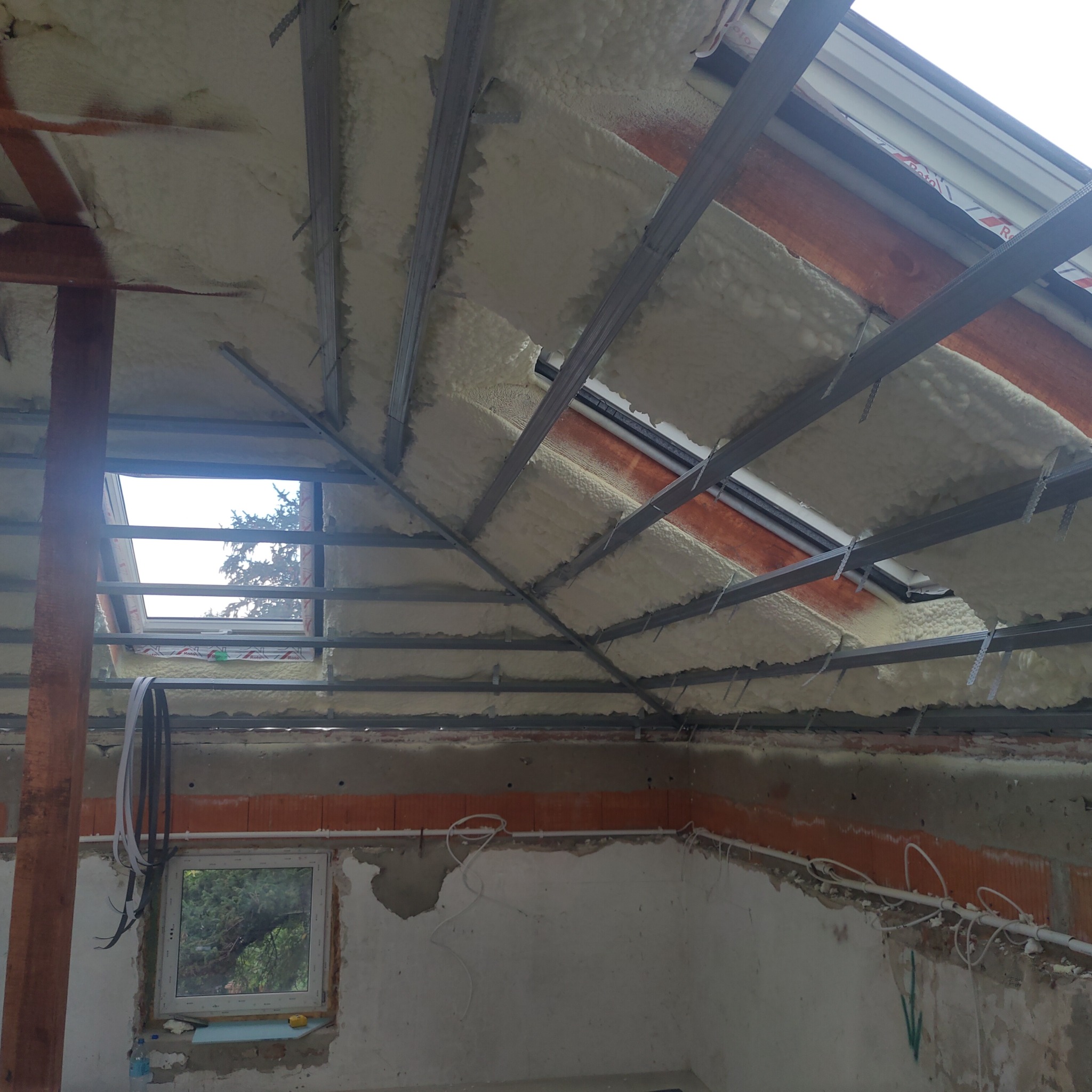 Poddasze z oknami dachowymi po ociepleniu pianą, widoczne metalowe profile konstrukcyjne i fragmenty instalacji elektrycznej na ścianach.