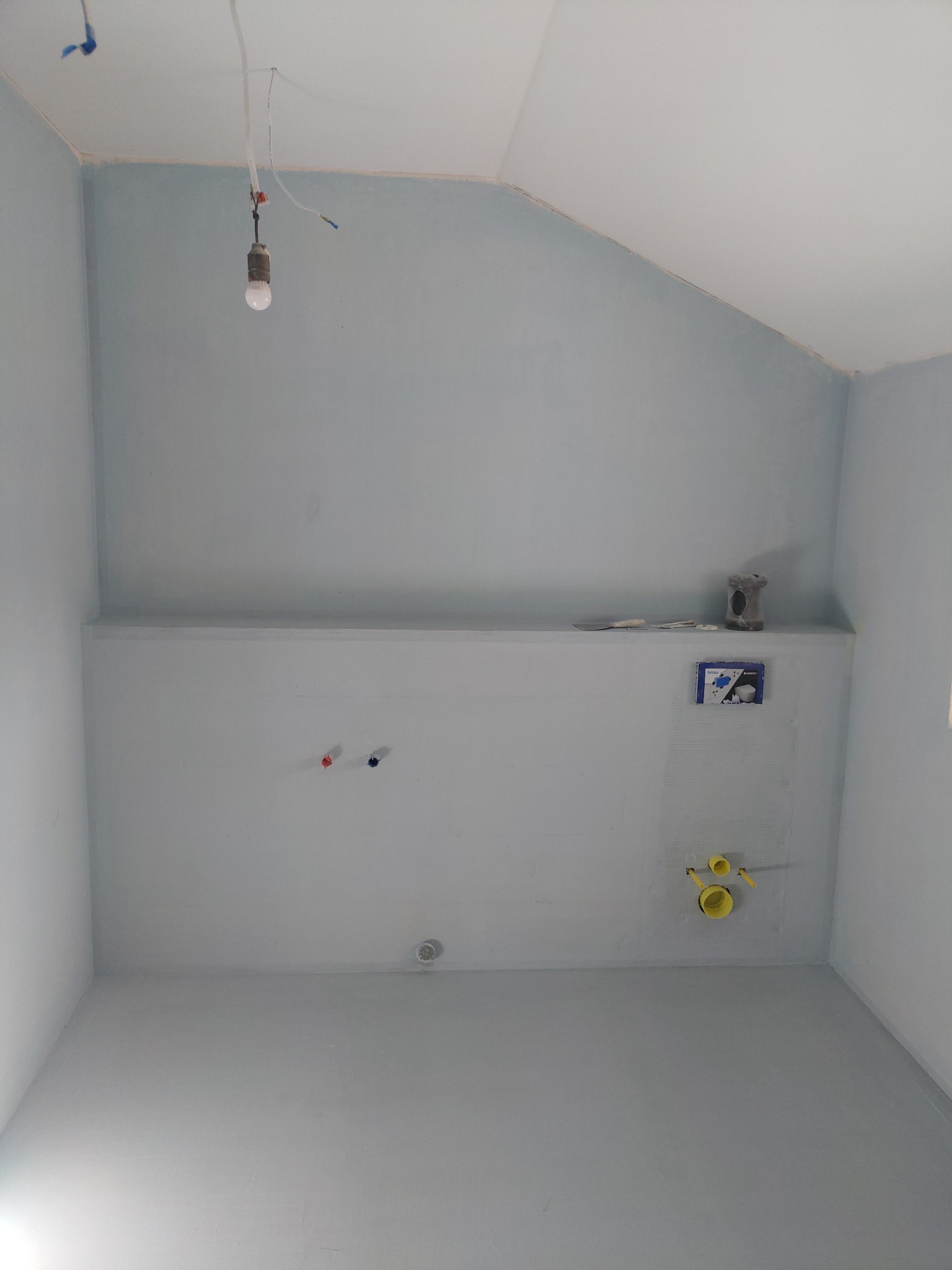 Wnętrze łazienki w trakcie remontu: pomalowane na szaro ściany i podłoga, widoczne instalacje wodno-kanalizacyjne, wisząca żarówka. Etap przygotowania do dalszych prac wykończeniowych.