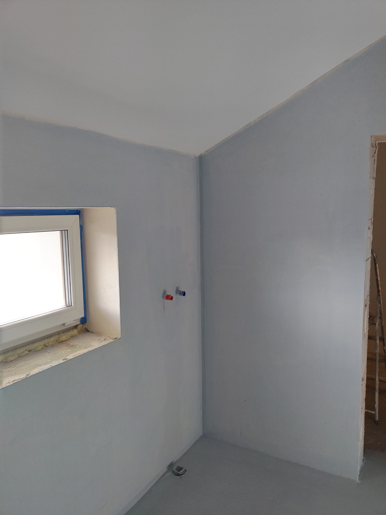 Szare wnętrze w trakcie remontu: ściany pomalowane na szaro, okno z niebieską taśmą malarską, instalacja wodna i drabina w tle. Etap prac malarskich.