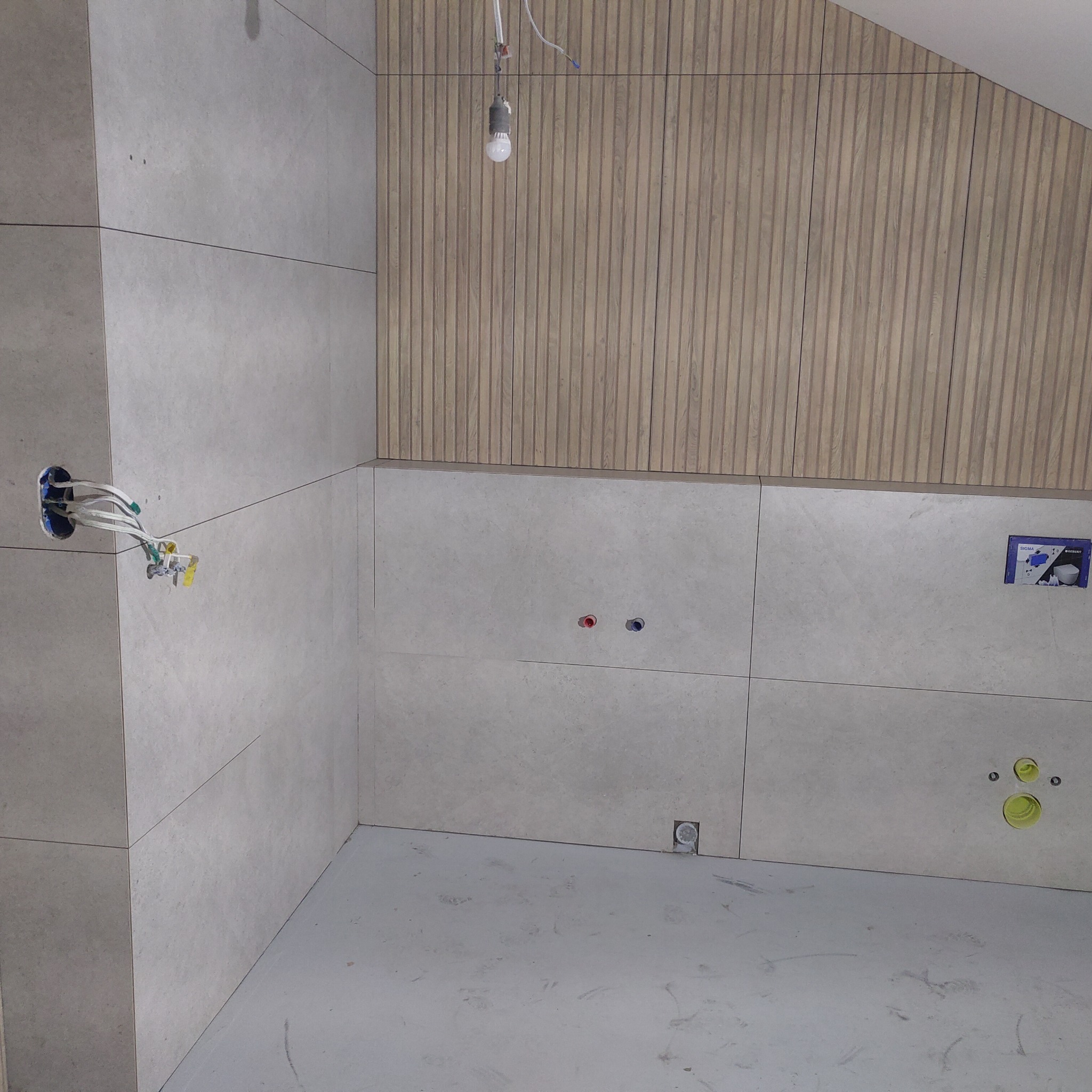 Łazienka w trakcie wykańczania: szare płytki na ścianach, drewniana okładzina, widoczne instalacje elektryczne i hydrauliczne, surowy beton na podłodze.