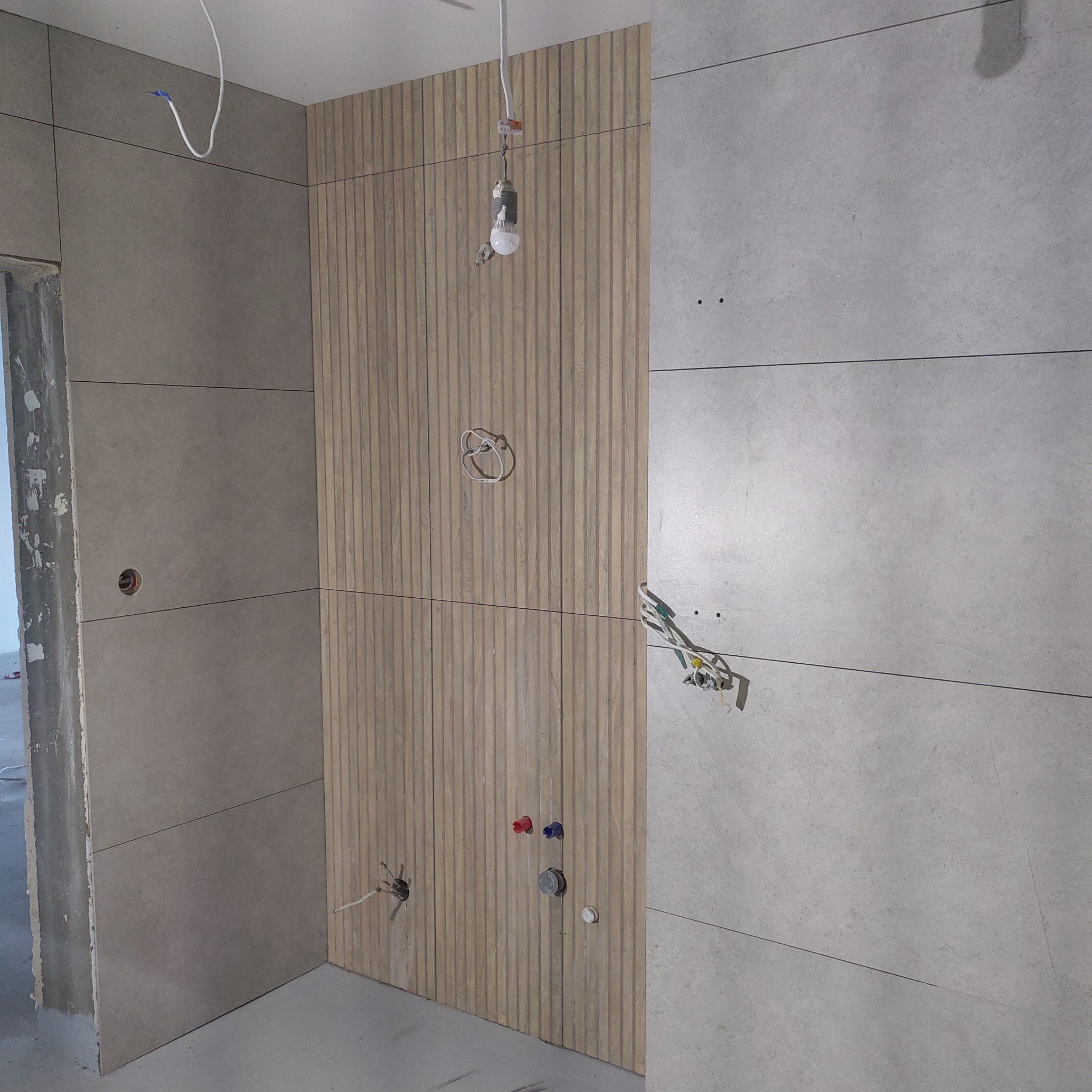 Ściana wyłożona szarymi płytami i panelami z drewnopodobnych lameli. Widoczne wystające przewody elektryczne i instalacja wodna. Stan surowy.
