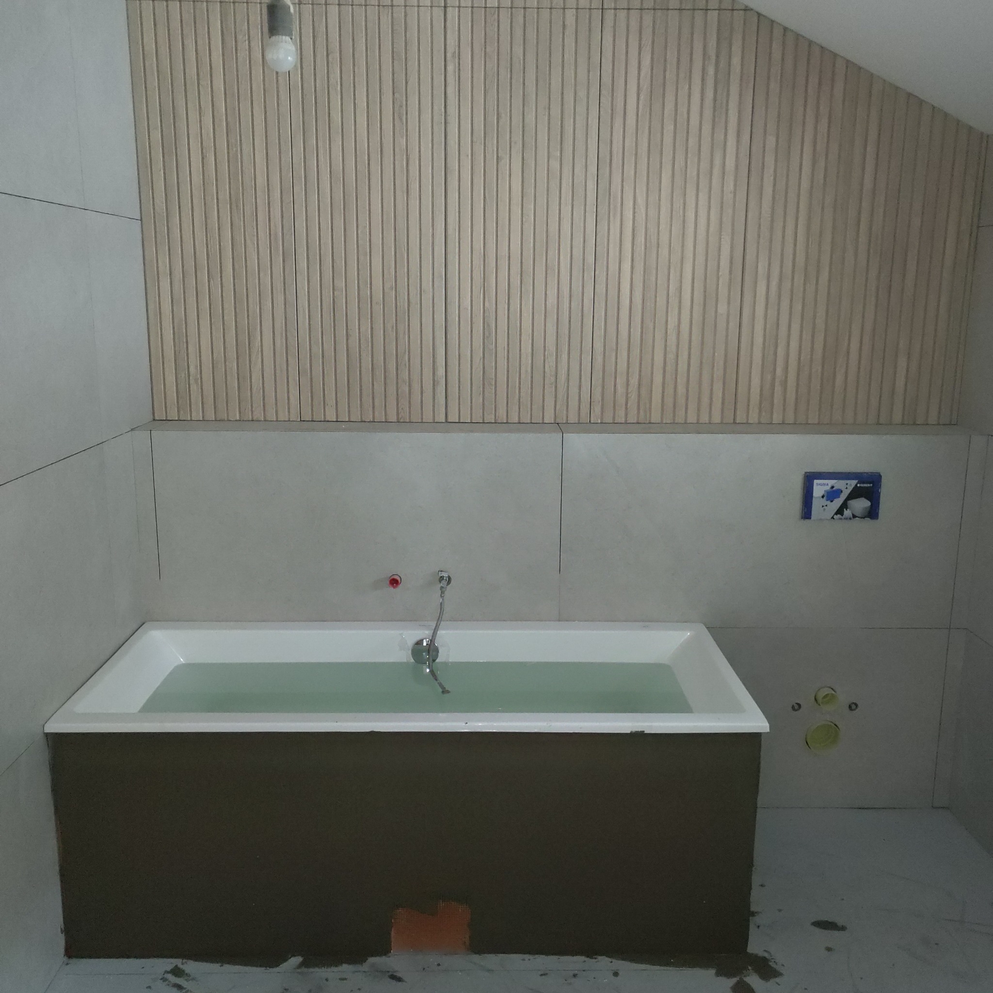 Wykończona łazienka w skandynawskim stylu: biała wanna z wodą, drewniane panele na ścianie i jasne płytki. Widoczne elementy instalacji wodnej i fragment podłogi.