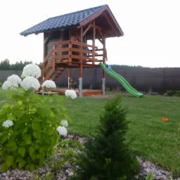 Drewniany plac zabaw z zadaszeniem i zjeżdżalnią na trawniku, widok z perspektywy ogrodu z kwitnącymi hortensjami na pierwszym planie.