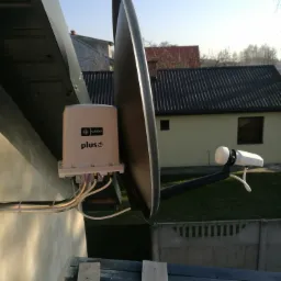 Zamontowana antena satelitarna Plus na elewacji budynku, widoczny konwerter i przewody, w tle dom z czarnym dachem.