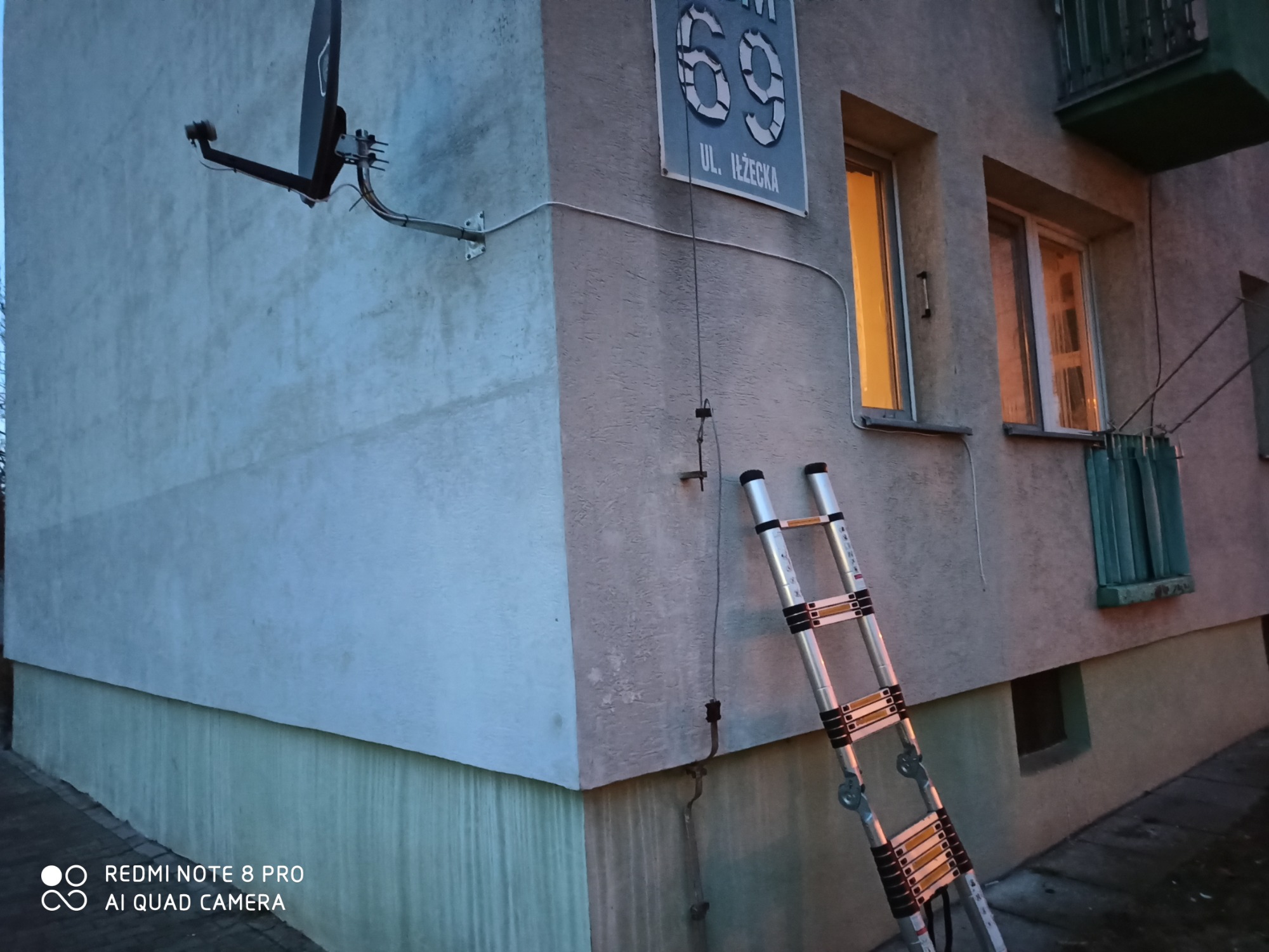 Antena satelitarna zamontowana na elewacji budynku, widoczna drabina oparta o ścianę, tabliczka z numerem budynku i nazwą ulicy.