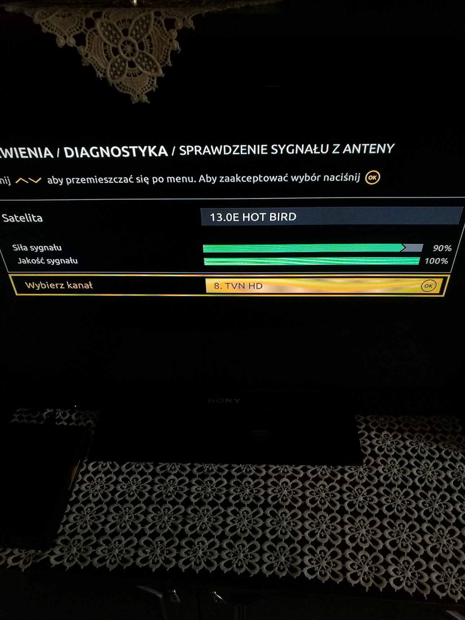 Ekran telewizora Sony wyświetlający menu diagnostyczne siły i jakości sygnału satelitarnego 13.0E Hot Bird z wartościami 90% i 100% w otoczeniu ciemnego wnętrza.