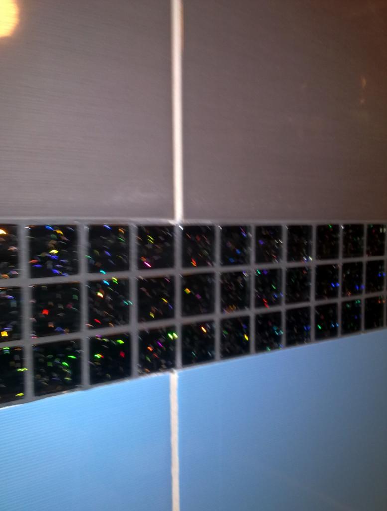 Fragment ściany z błyszczącą mozaiką między szarymi i błękitnymi płytkami, widok z bliska pod kątem.