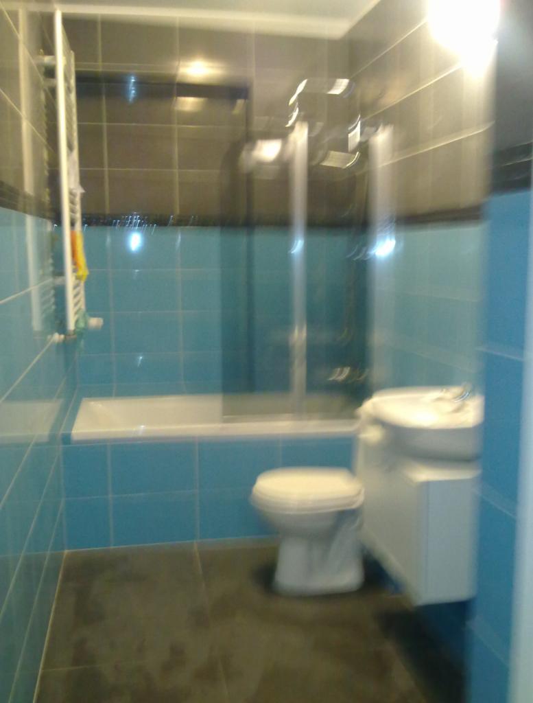 Niebiesko-szara łazienka z wanną za szklanym parawanem, umywalką z szafką i toaletą, widoczne płytki na ścianach i ciemna podłoga.