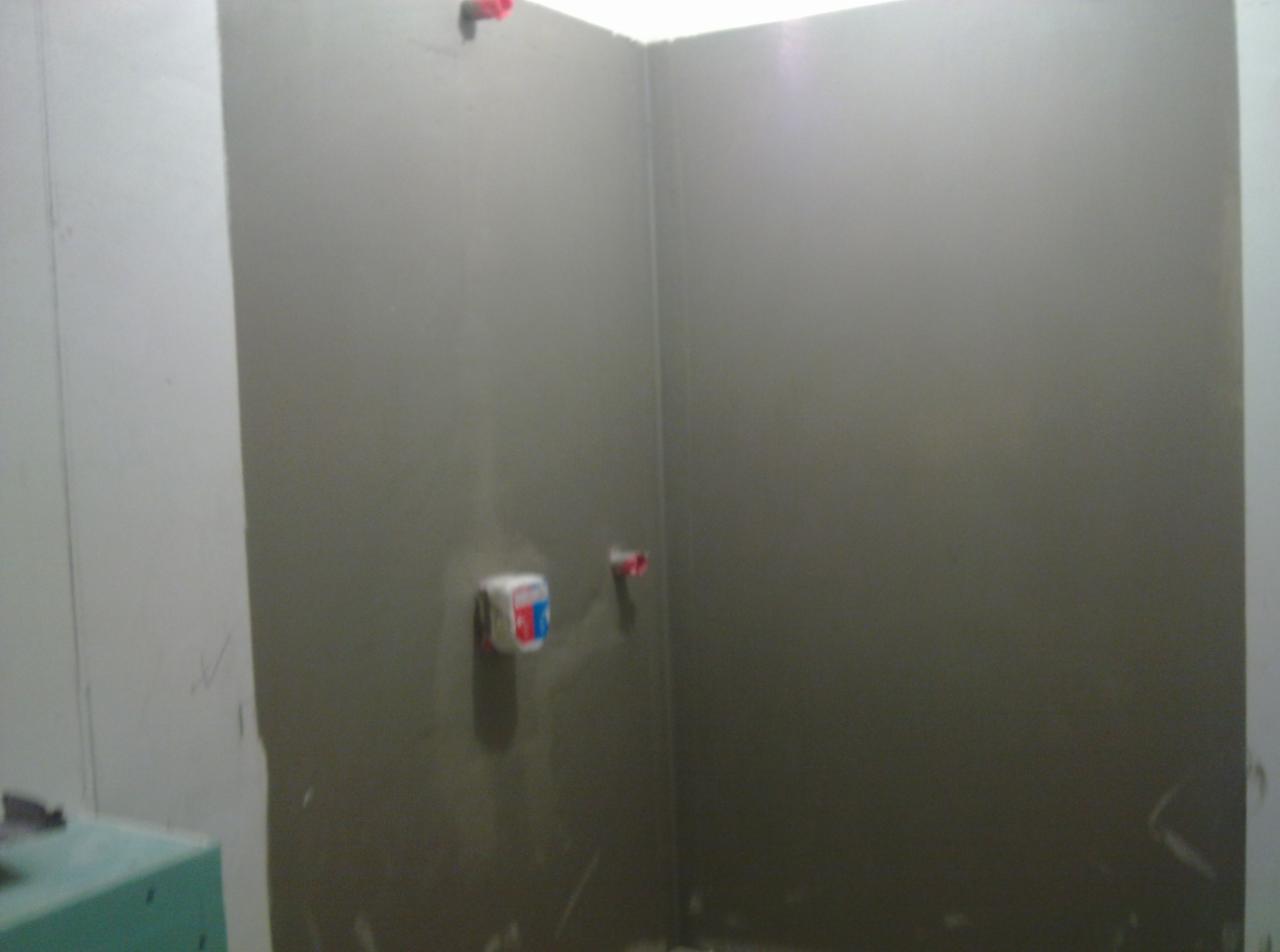 Przygotowanie ścian w narożniku łazienki pod płytki, widoczne zamontowane puszki pod baterie i zaklejone rury, ściany pokryte szarą warstwą wyrównującą.