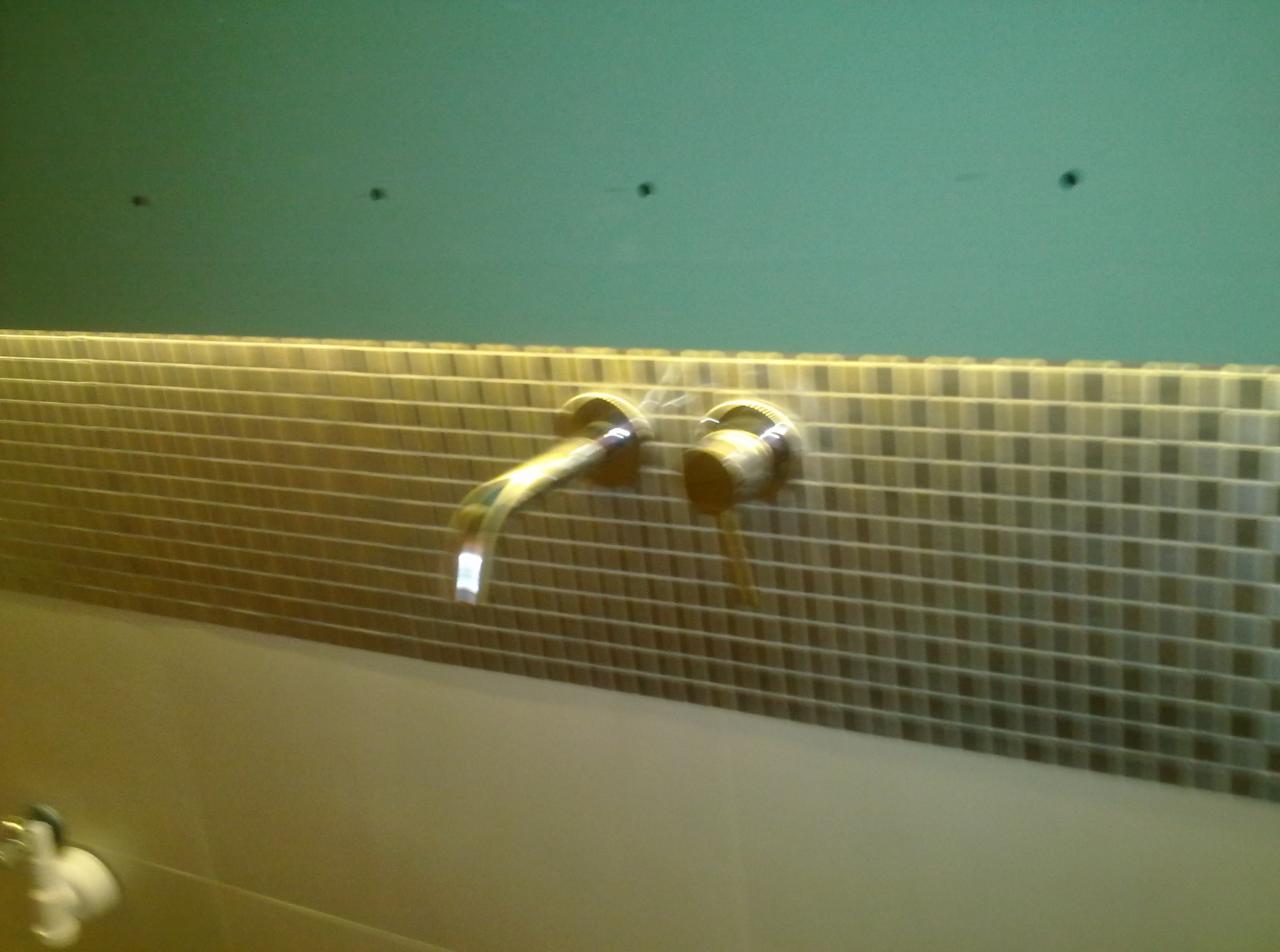 Bateria umywalkowa podtynkowa, chromowana, zamontowana na ścianie wyłożonej mozaiką w odcieniach beżu i brązu, z widocznymi otworami w ścianie powyżej.