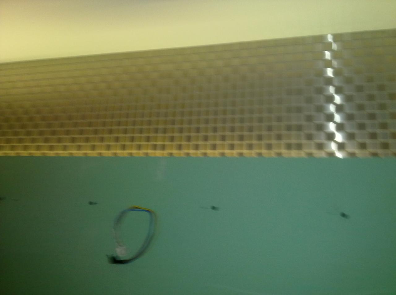 Niedokończona ściana z zielonej płyty gipsowo-kartonowej z widocznymi przewodami i metalową siatką na górze, w tle jasny sufit.