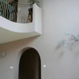 Wnętrze domu z wysokim sufitem, łukowatym przejściem i balkonem z metalową balustradą, na którym stoi doniczka z zieloną rośliną. Ściany pomalowane na jasny, neutralny kolor, oświetlone przez...