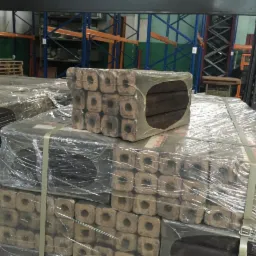 Ułożone na palecie kostki pelletu drzewnego, zapakowane w folię i zabezpieczone taśmą, widoczne w magazynie z regałami w tle.