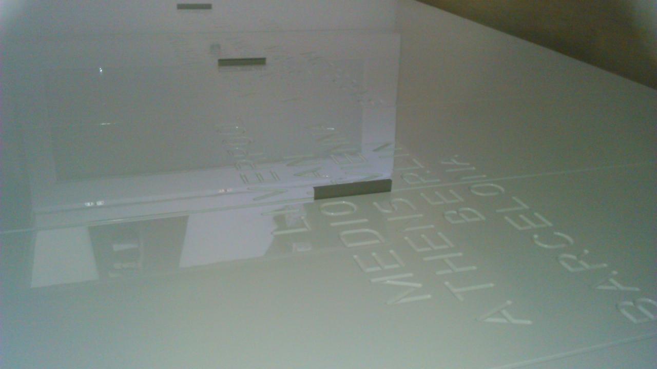 Szklana ściana działowa z wygrawerowanymi nazwami miast, oświetlona z góry, widziana pod kątem, z widocznymi elementami mocującymi.