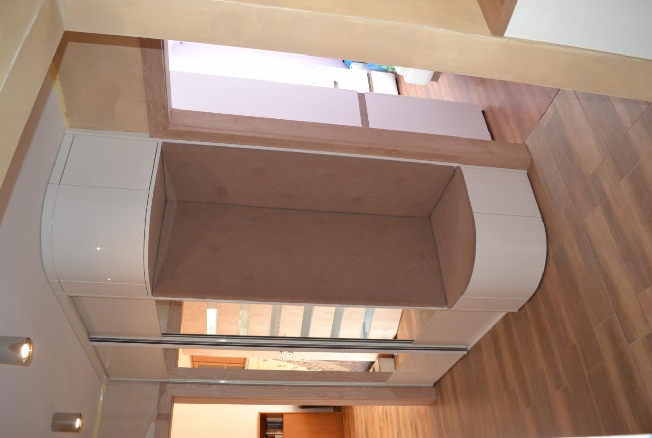 Wnęka z siedziskiem obita beżową tkaniną, wbudowana w szafę z przesuwnymi drzwiami i zaokrąglonymi bokami, widok z dołu na jasną podłogę.