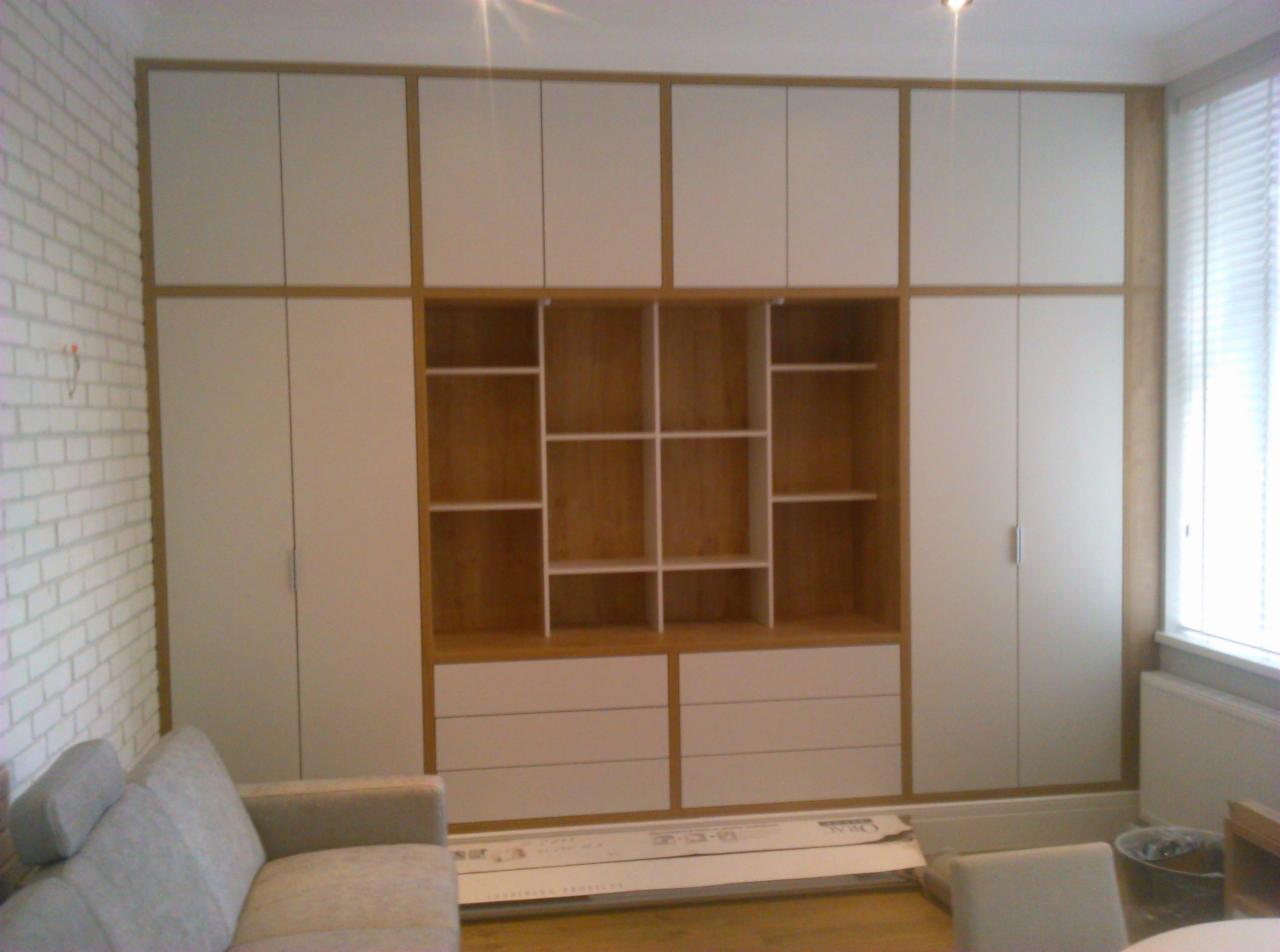 Biała szafa wnękowa z drewnianymi akcentami, z otwartymi półkami i szufladami, częściowo zasłonięta przez sofę w szarym odcieniu, na tle ściany z białej cegły.