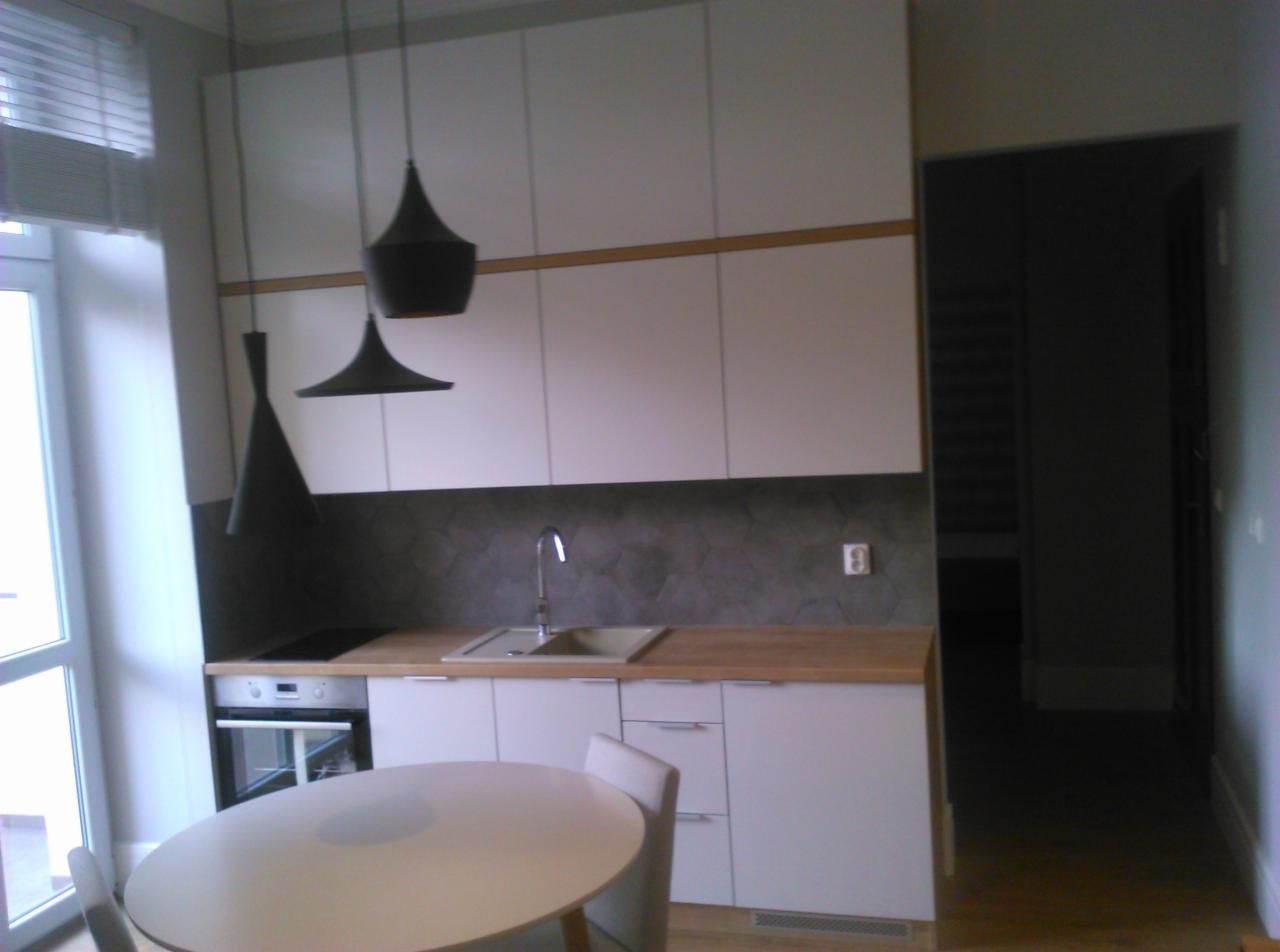 Nowoczesna kuchnia z białymi szafkami, drewnianym blatem i heksagonalnymi płytkami na ścianie, oświetlona wiszącymi lampami w kształcie stożków i okrągłym stołem na pierwszym planie.