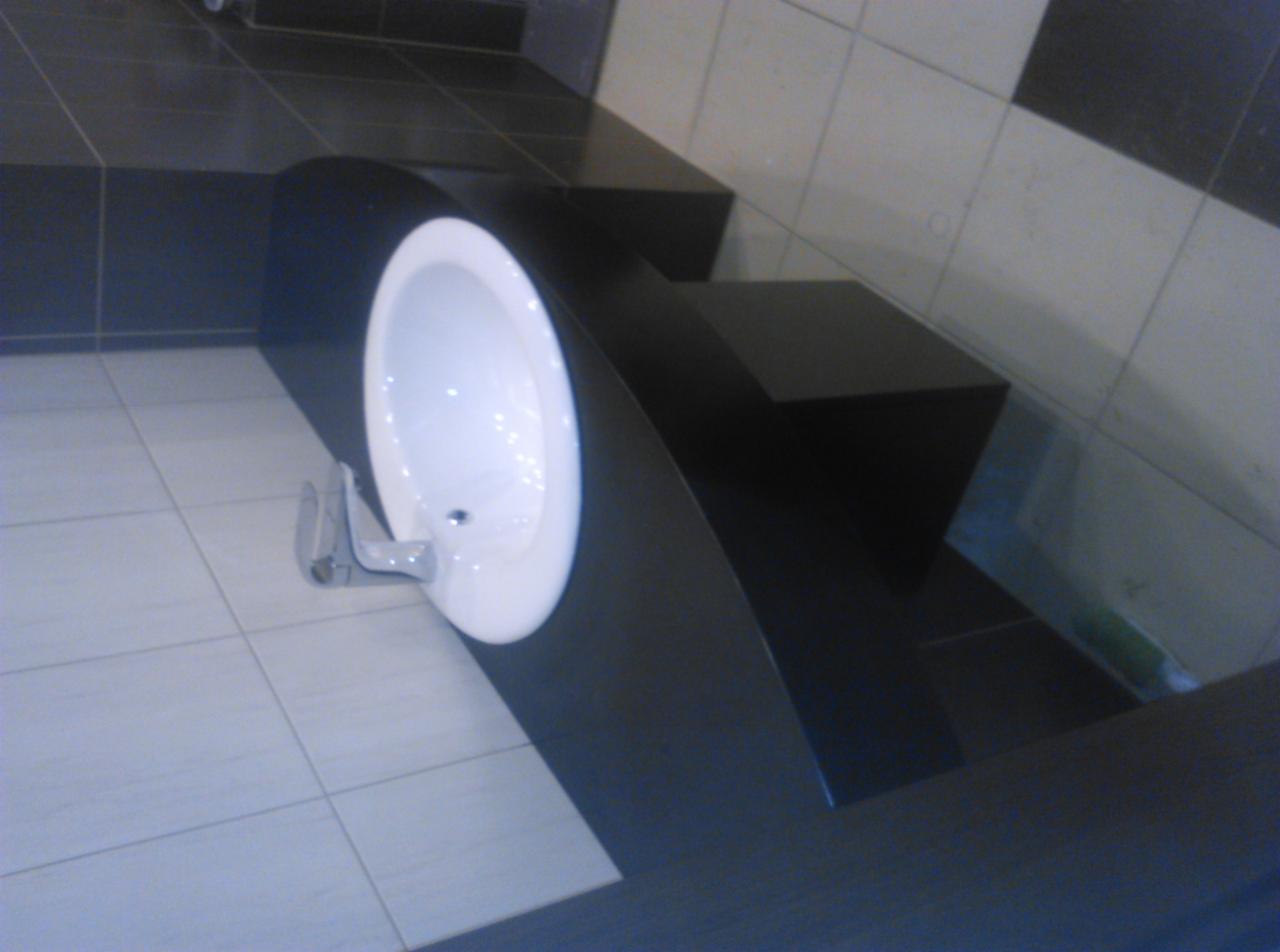 Umywalka wpuszczona w czarny, niestandardowy blat o zaokrąglonym kształcie, z chromowaną baterią, na tle biało-czarnych płytek podłogowych i ściennych.