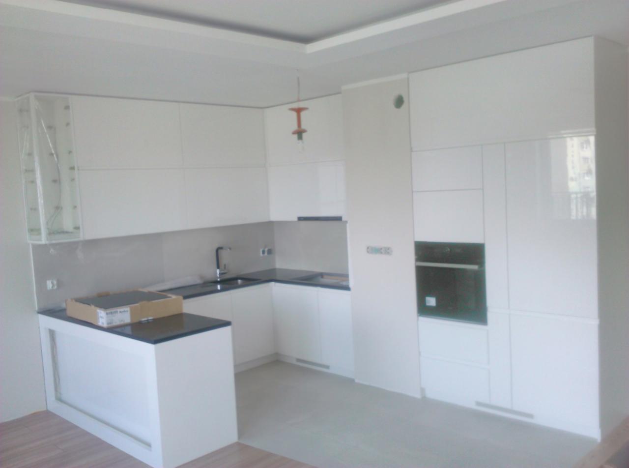 Nowoczesna, biała kuchnia z połyskiem, czarnym blatem i wbudowanym piekarnikiem, częściowo jeszcze zabezpieczona folią ochronną, z kartonem na blacie, w trakcie wykańczania.