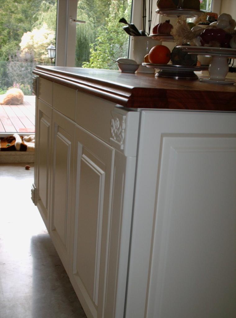 Biała wyspa kuchenna z ciemnym, drewnianym blatem, zdobionymi frontami szafek i dekoracyjnymi elementami, widok z boku, na blacie patery z owocami i akcesoriami kuchennymi, w tle widoczny taras...