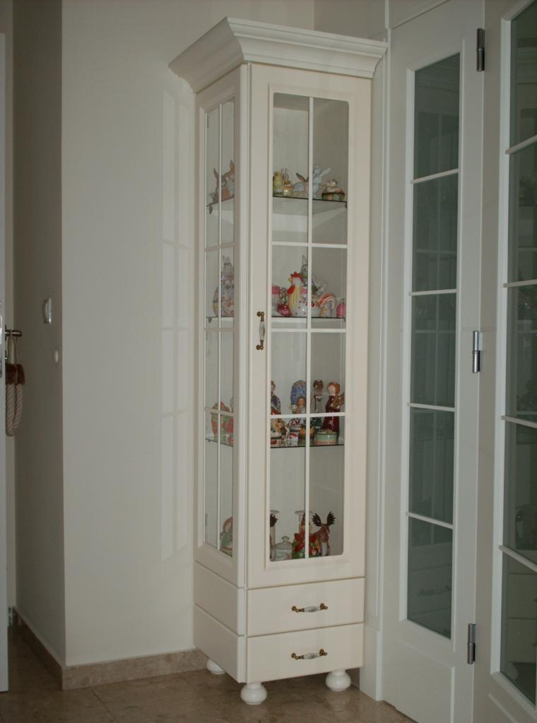 Wysoka, wąska, przeszklona witryna w kolorze kremowym, stojąca w rogu pokoju w Brzozie, wypełniona kolekcją figurek i ozdobnych przedmiotów.