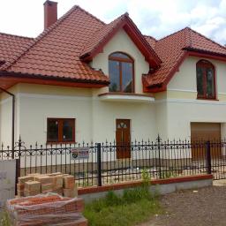 Usługi Remontowo-budowlane BUDMAX - Nowo wybudowany dom jednorodzinny z jasną elewacją, czerwoną dachówką i czarnym, kutym ogrodzeniem, z paletą cegieł na pierwszym planie, sugerujący prace wykończeniowe.