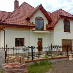 Nowo wybudowany dom jednorodzinny z jasną elewacją, czerwoną dachówką i czarnym, kutym ogrodzeniem, z paletą cegieł na pierwszym planie, sugerujący prace wykończeniowe.