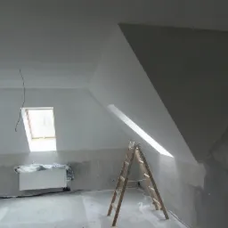 Poddasze w trakcie remontu: drewniana drabina oparta o ścianę przy oknie dachowym, widoczny grzejnik pod oknem na ścianie szczytowej, ściany częściowo przygotowane do malowania.