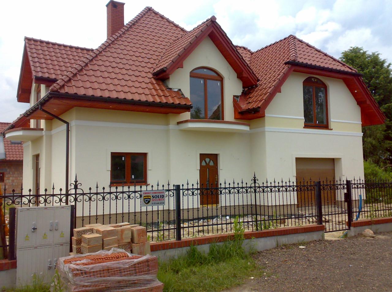 Nowo wybudowany dom jednorodzinny z jasną elewacją, czerwoną dachówką i czarnym, kutym ogrodzeniem, z paletą cegieł na pierwszym planie, sugerujący prace wykończeniowe.