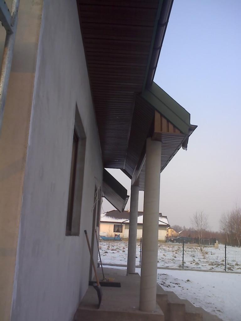 Elewacja domu w trakcie ocieplania, widoczna podbitka dachowa drewniana, szara ściana i kolumny, na ziemi leży łopata i miotła, w tle zaśnieżony krajobraz.