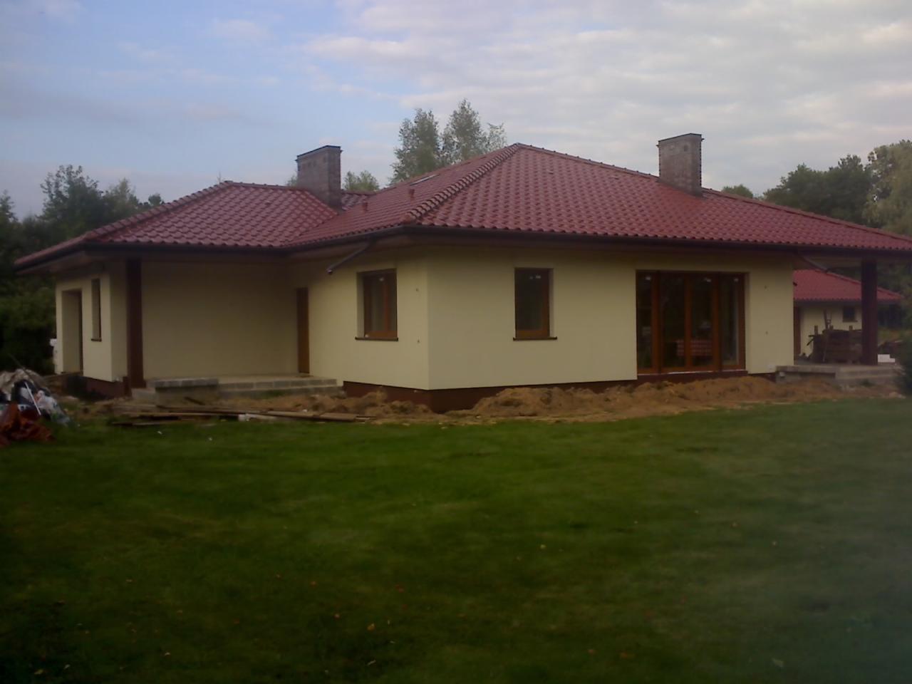 Jasny, parterowy dom z czerwoną dachówką i brązowymi ramami okiennymi, otoczony trawnikiem i fragmentami ziemi po pracach budowlanych. Widoczny fragment drugiego budynku w tle.