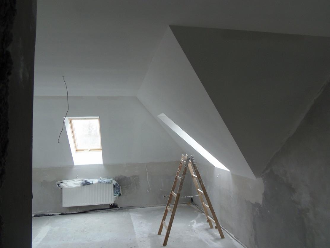 Poddasze w trakcie remontu: drewniana drabina oparta o ścianę przy oknie dachowym, widoczny grzejnik pod oknem na ścianie szczytowej, ściany częściowo przygotowane do malowania.