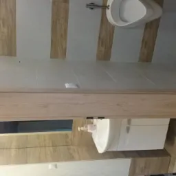 Wykończona łazienka z beżowo-białymi płytkami, drewnianą ramą drzwi i białym umywalką z szafką. Widoczne elementy armatury i dozownik mydła. Neutralna kolorystyka.