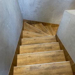 P.W.OPTIVA Marcin Dobosz - Kątowe ujęcie schodów wyłożonych panelami imitującymi drewno z białymi listwami, na tle szarej ściany o fakturze betonu. Widok z góry.
