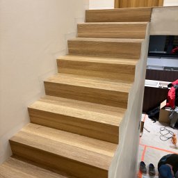 P.W.OPTIVA Marcin Dobosz - Dębowe schody z widocznymi słojami drewna, zamontowane na betonowej konstrukcji. W tle widoczne narzędzia i elementy wykończeniowe, sugerujące trwający montaż.