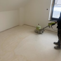 P.W.OPTIVA Marcin Dobosz - Szlifowanie wylewki samopoziomującej w pokoju na poddaszu. Widoczna maszyna do szlifowania i częściowo osoba obsługująca. Ślady po szlifowaniu na jasnej powierzchni.