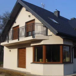 Dwupiętrowy dom z balkonem w Piastowie. Kremowa elewacja, ciemna dachówka, okna z brązowymi roletami. Widok z poziomu gruntu, boczny.