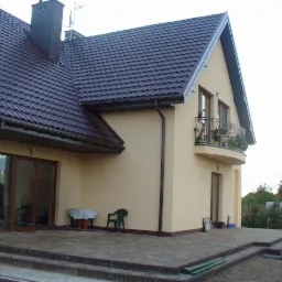 Elewacja domu jednorodzinnego z balkonem, brązowy dach z ceglanym kominem, jasny tynk. Widoczne elementy wykończenia tarasu i ogrodu. Ujęcie z dołu, słoneczny dzień.