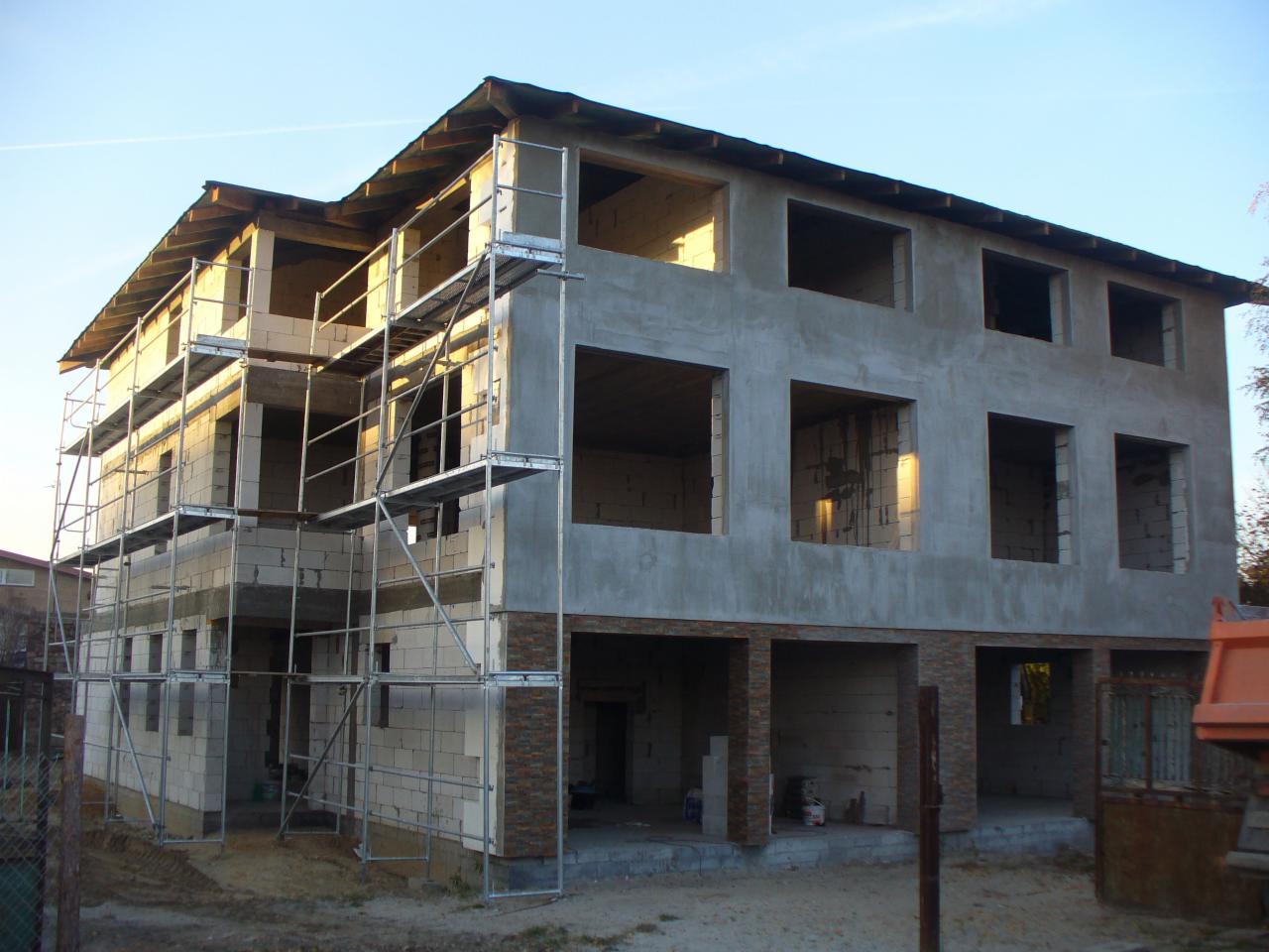 Surowy, dwukondygnacyjny dom w trakcie budowy, z widocznymi rusztowaniami, oknami bez szyb i częściowo otynkowaną elewacją. Widoczne bloki i kolumny z cegły.